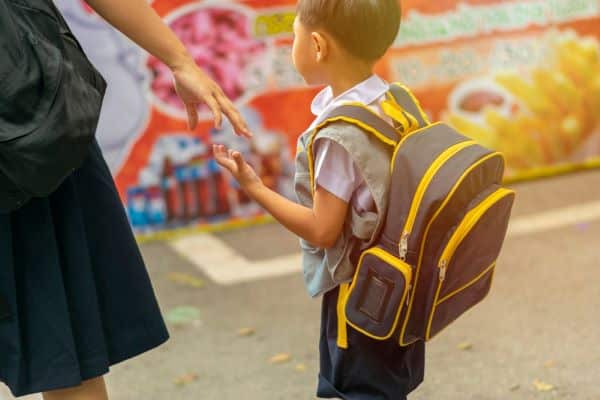 Irmâ mais velha segura a mão do menininho que veste uniforme escolar e usa uma mochila nas costas. A volta às aulas e a rotina escolar.