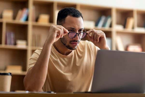 Homem negro, jovem, demonstra incômodo visual tentando regular os óculos frente ao notebook. Um sintoma das condições oculares astigmatismo e hipermetropia. condições