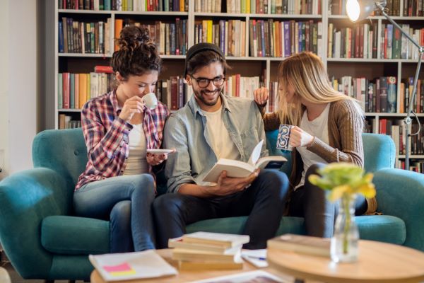 Três jovens leem livro  sentados em um sofá num ambiente com boa iluminação - Conceito de ler e seus mitos.