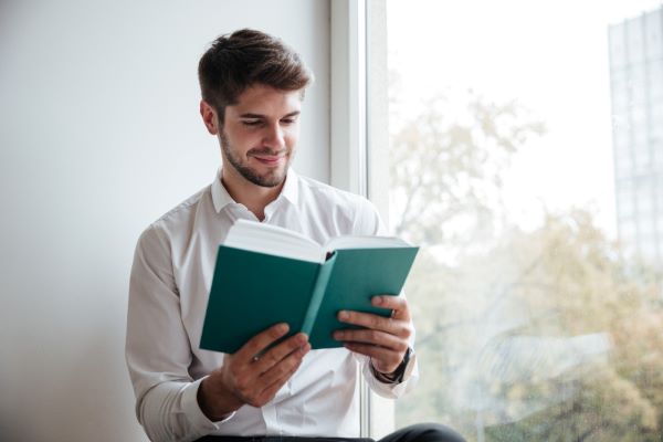 Homem jovem lê próximo a uma janela, em condições ideais de iluminação. O hábito de ler e seus cuidados.