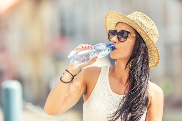 Jovem bebe água e se protege do sol usando óculos e chapéu; formas de se evitar a fotossensibilidade.