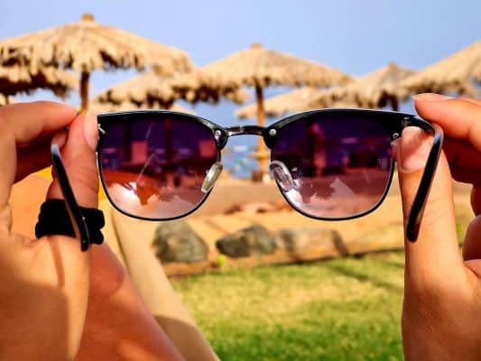 Jovem segura entre as mãos um par de óculos de sol, demonstrando a importância de seu uso durante os meses quentes do verão, especialmente em locais como a praia.
