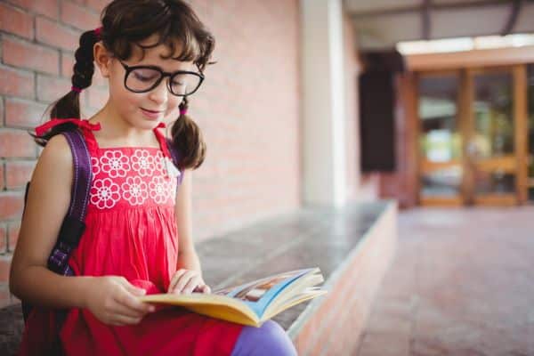 Menininha, usando óculos pretos e vestindo uma blusinha vermelha com flores brancas, lê um livro de histórias sentada no pátio da escola enquanto aguarda a volta às aulas.