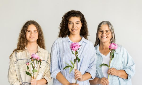 Três gerações de mulheres, avó, mãe e filha, sorridentes, seguram uma rosa recebida pela homenagem ao Dia da Mulher.