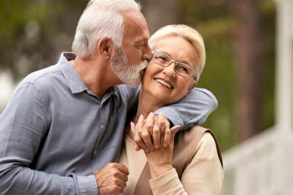 Casal de idosos felizes passeando pelas ruas. A senhora está recebendo um beijo do companheiro. 
A idade avançada é um dos riscos de desenvolver degeneração macular relacionada à idade.