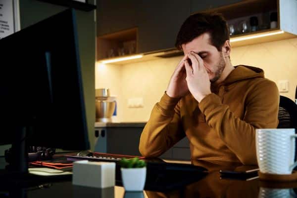 Homem jovem, vestindo moletom marrom, tem as mãos sobre os olhos indicando sinal de desconforto ocular em razão do uso de computador em ambiente climatizado no inverno. 