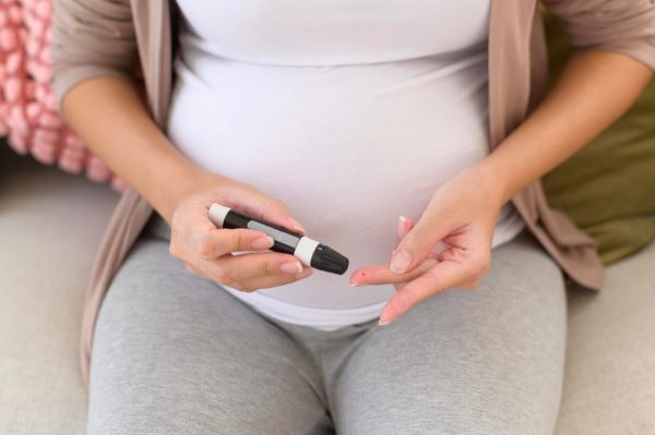 Gestante sentada no sofá, vestindo legging cinza, camiseta branca e casaquinho rosado mede os níveis de açúcar no sangue. Controlando a diabete gestacional.