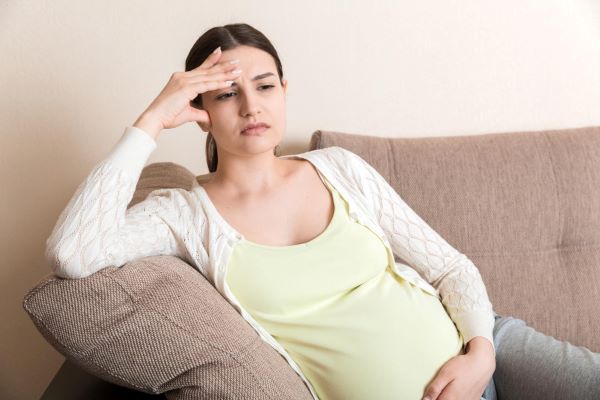 Mulher jovem, grávida, vestindo uma camiseta amarelinha e um casaquinho branco está apoiada no braço do sofá demonstrando desconforto. Preocupa-se com a retinopatia diabética.