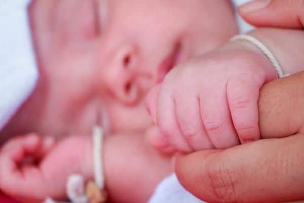 Close da mãozinha de um bebê recém nascido segurando o dedo de um adulto. Observa-se seu rostinho e o o outro bracinho em imagem desfocada. Conceito de retinopatia da prematuridade.