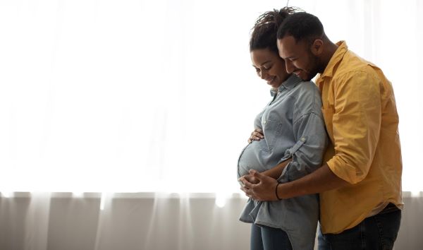 Casal de jovens negros abraçados acariciam a barriga da gestante. Estão frente a uma cortina branca de voal. Ela veste uma camisa jeans e ele uma amarela.