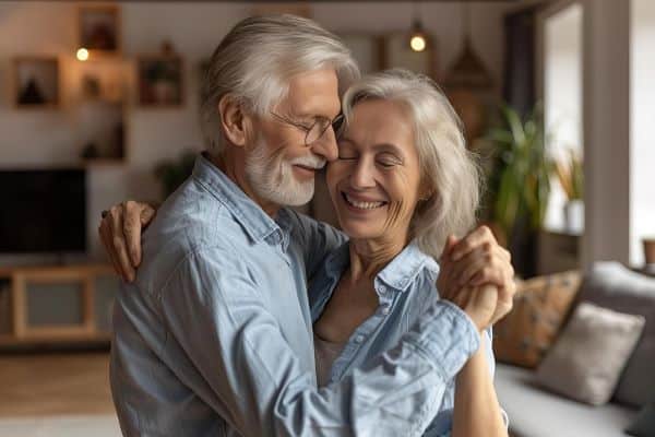 Casal de idosos, sorridentes, dançam juntinhos na sala de sua casa. Ambos vestem camisas jeans.