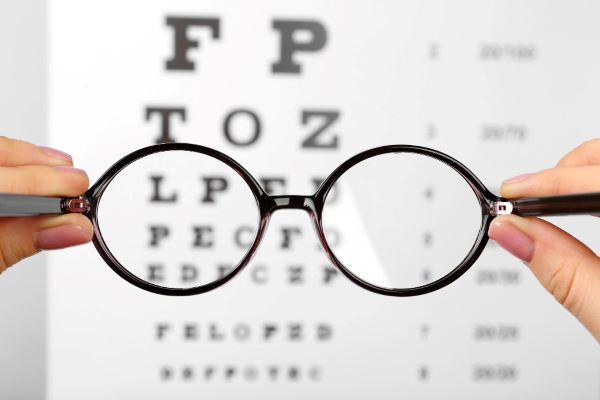 Mãos femininas seguram um par de óculos de grau, preto, que está frente à tabela oftalmológica. A imagem das letras, através das lentes, está borrada. Conceito de miopia.