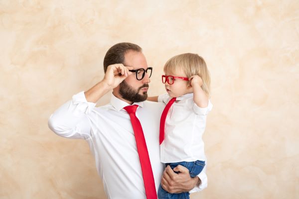 Pai segurando o filho pequeno nos braços. Ambos divertem-se mexendo em seus óculos. Vestem roupas iguais: camisa branca, gravata vermelha e calça jeans.