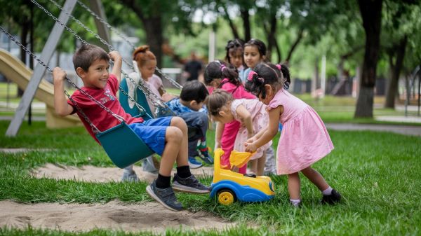 Grupo de crianças pequenas brinca em um parquinho com brinquedos coloridos e em balanços.