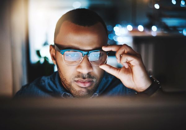 Homem negro, ajeitando os óculos de grau com filtro de luz azul em seu rosto, trabalha frente a um computador. 