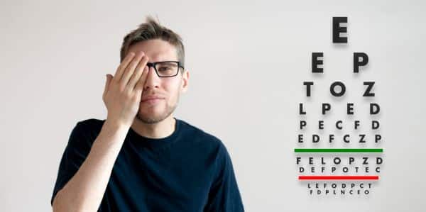 Jovem com barba aparada, cabelo curto e claro, vestindo uma camiseta preta e usando óculos pretos, verifica a visão por meio de exame ocular com o gráfico de letras de teste.