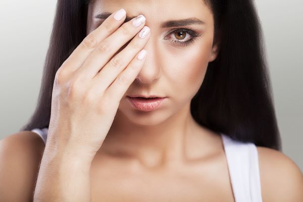 Jovem com olhos castanhos, cabelos pretos e lisos tapa o olho direito com uma das mãos em sinal de desconforto.