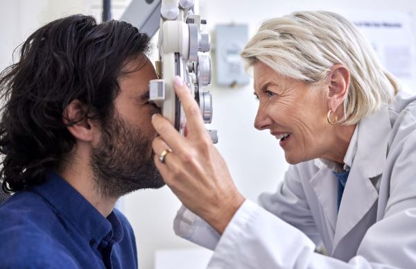Médica, mulher madura com cabelos grisalhos, vestindo jaleco branco, examina os olhos de um paciente jovem, que usa barba e tem cabelos longos e escuros.
