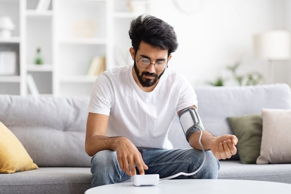 Jovem com cabelos e barba escuros, usando óculos de grau, mede a pressão com um aparelho.