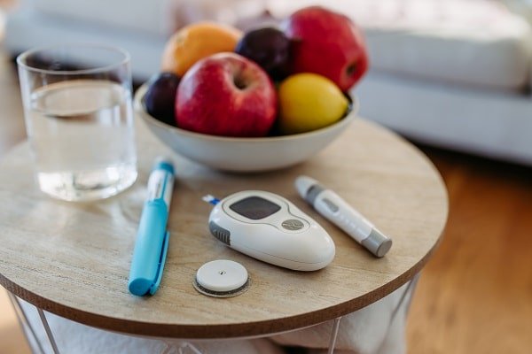 Sobre uma mesinha de madeira, um recipiente com frutas, um copo de água, caneta de insulina e instrumentos de medir glicose no sangue.