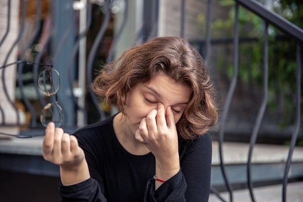 Mulher jovem sentada numa escadaria, segura um par de óculos na mão direita enquanto esfrega os olhos com a outra mão.