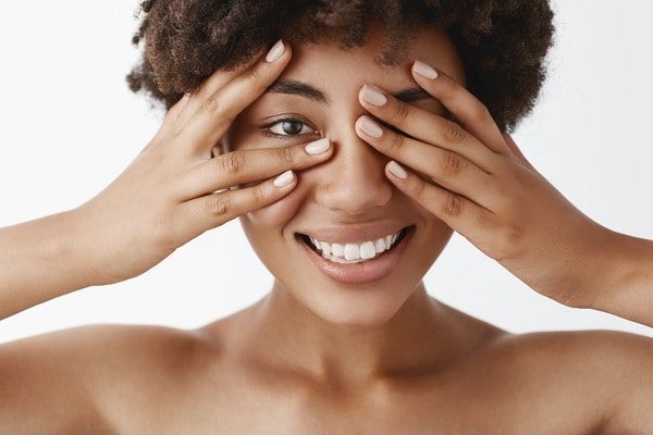 Linda mulher negra, jovem, tapando parcialmente os olhos com as mãos e sorrindo.
