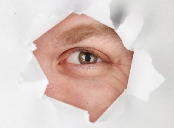 Olho masculino visto através de um rasgo num papel branco que cobre o rosto do homem.